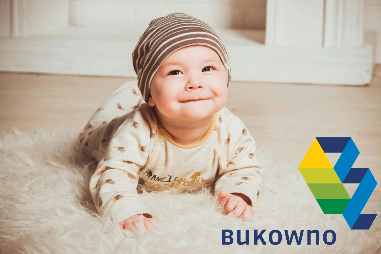 Zdjęcie przedstawia uśmiechnięte niemowlę leżące na brzuszku na miękkim, białym dywanie z długim włosiem (typu futrzak).Szczegóły wyglądu dzieckaDziecko ma uniesioną wysoko głowę i klatkę piersiową, opierając się stabilnie na przedramionach.Patrzy przed siebie z radosnym, łagodnym uśmiechem.Na głowie ma ubraną bawełnianą czapkę w poziome, brązowo-białe paski, która zakrywa uszy.Ubrane jest w jasny pajacyk lub piżamkę we wzory (widoczne są m.in. motywy przypominające żołędzie lub listki) oraz napis na piersi „Francis”.Tło i elementy graficzneTło za dzieckiem jest jasne, rozmyte, z widoczną drewnianą podłogą oraz jasnymi elementami mebli lub ściany.W prawym dolnym rogu zdjęcia umieszczono duże logo przedstawiające geometryczną, warstwową literę „B” lub wstęgę w kolorach żółtym, zielonym i niebieskim.Pod logo znajduje się wyraźny, granatowy napis „Bukowno”.
